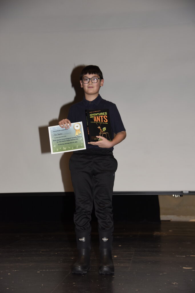 A young boy stands on stage holding up a certificate and a book titled "Adventures Among Ants".